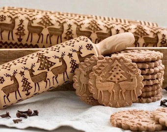 以下が含まれることがあります： トナカイとクリスマスツリーが描かれたクリスマスをテーマにしたデザインのウッドローリングピン。ローリングピンは、クッキーに装飾的なパターンを作成するために使用されます。同じデザインのクッキーの積み重ねが画像に表示されています。