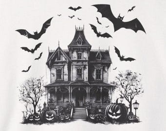 May include: A cream-coloured sweatshirt featuring a black graphic print of a haunted Victorian-style house. The scene includes several carved jack-o'-lanterns, bare trees, and a flock of bats flying above the house against a plain background.