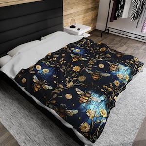 Pode incluir: Um cobertor azul escuro com um padrão repetido de abelhas, flores e folhagens em tons de ouro e amarelo. O design tem um efeito de vitral com destaques azuis e amarelos. O cobertor está em uma cama com lençóis brancos.