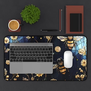 May include: A desk mat with a bee and floral design, a laptop, and a white mouse. The mat features a dark blue background with gold and white accents. A cup of coffee, a small plant, and stationery are also on the dark surface.