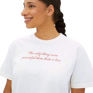 May include: White t-shirt with the red text, "The only thing more powerful than hate is love." The shirt has short sleeves and a crew neck. The text is in a cursive font.
