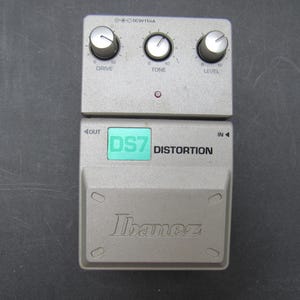 May include: A silver Ibanez DS7 Distortion guitar effects pedal featuring three control knobs labeled Drive, Tone, and Level. The unit includes a green square label with the text DS7, a small red indicator light, and input and output ports.