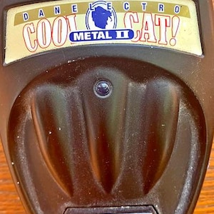 May include: A dark brown Danelectro Cool Cat Metal II guitar effects pedal featuring three top-mounted control knobs labeled vol, switch (hi lo off), and dist. The central faceplate displays the brand name, a cat logo, and a small indicator light.