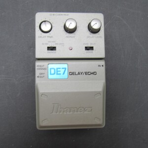 May include: A grey Ibanez DE7 Delay/Echo guitar effects pedal featuring three control knobs for delay time, repeat, and delay level. The unit includes two toggle switches for range and mode, a small indicator light, and a large foot switch.