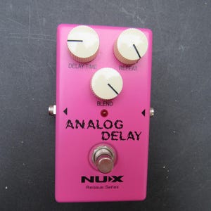 May include: A bright pink NUX Analog Delay guitar effects pedal featuring three cream-colored control knobs labeled Delay Time, Repeat, and Blend. The pedal includes a central metal footswitch, a small red indicator light, and black text.