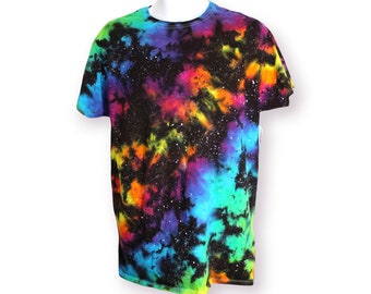 Könnte beinhalten: Ein kurzärmeliges T-Shirt mit einem lebendigen Batik-Design, das einer Galaxie ähnelt. Das Shirt hat eine schwarze Basis mit Farbspritzern in Blau, Grün, Gelb, Orange und Lila sowie weißen Sprenkeln. Das Shirt wird auf einer weißen Schaufensterpuppe präsentiert.