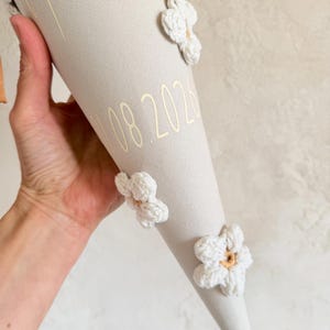 May include: A beige fabric cone decorated with three white crocheted flowers featuring tan centers. The cone displays the name Malina in gold script lettering and the date 10.08.2026 in gold numerals. A brown ribbon is visible at the top edge.
