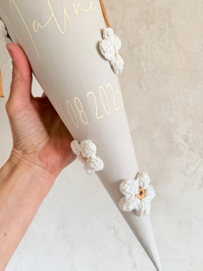 May include: A beige fabric cone decorated with three white crocheted flowers featuring tan centers. The cone displays the name Malina in gold script lettering and the date 10.08.2026 in gold numerals. A brown ribbon is visible at the top edge.