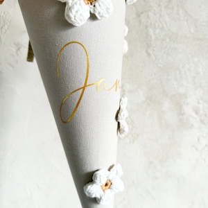 May include: A light gray cone-shaped fabric gift container decorated with white crocheted flowers featuring tan centers. The name Jan is printed in gold cursive script on the front. A gold glittery ribbon trim is visible at the top edge.