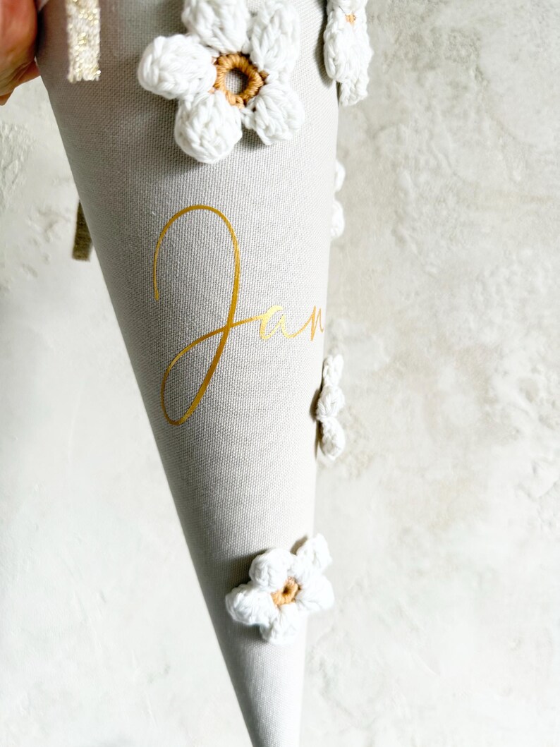 May include: A light gray cone-shaped fabric gift container decorated with white crocheted flowers featuring tan centers. The name Jan is printed in gold cursive script on the front. A gold glittery ribbon trim is visible at the top edge.