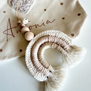 May include: A cream-colored canvas bag with gold polka dots and the name "Antonia" in brown script. A handmade rainbow-shaped wall hanging in cream and tan tones is in the foreground. A wooden bead garland with a bell hangs from the bag.