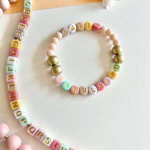 Pu&ograve; includere: Un braccialetto e un portachiavi con perline color pastello con scritte e dettagli dorati. Il braccialetto recita "MALI SCHULKIND" con un mix di perline pastello. Il portachiavi presenta perline alfabetiche e perline di legno.