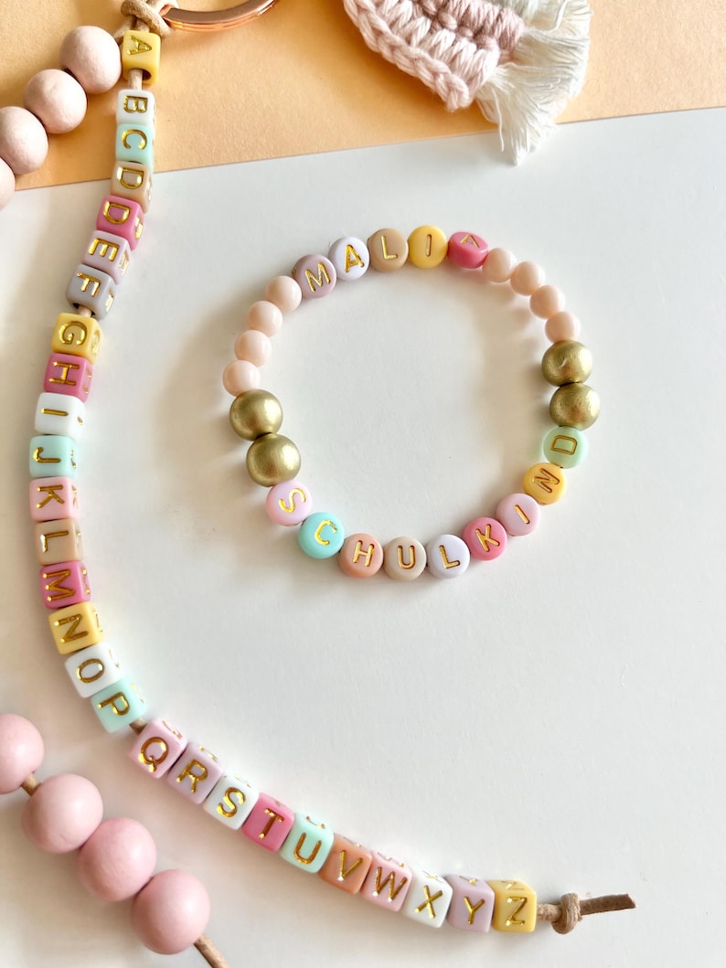 Pu&ograve; includere: Un braccialetto e un portachiavi con perline color pastello con scritte e dettagli dorati. Il braccialetto recita "MALI SCHULKIND" con un mix di perline pastello. Il portachiavi presenta perline alfabetiche e perline di legno.