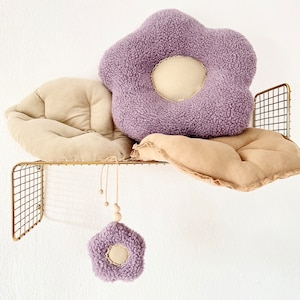 May include: Two plush flower-shaped cushions in beige and purple, with a smaller purple flower-shaped cushion hanging from a wooden bead garland. The cushions are displayed on a gold wire shelf.