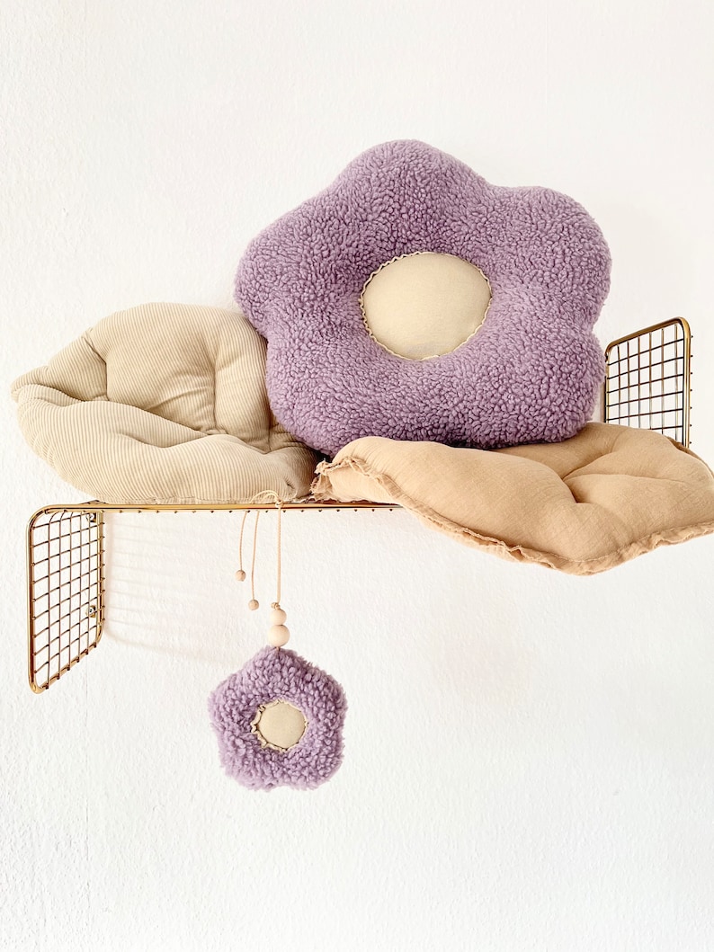 May include: Two plush flower-shaped cushions in beige and purple, with a smaller purple flower-shaped cushion hanging from a wooden bead garland. The cushions are displayed on a gold wire shelf.