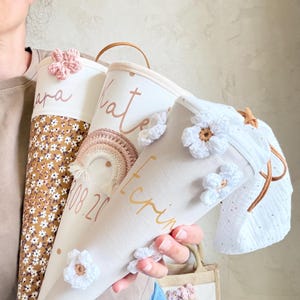 May include: Three cone-shaped fabric gift bags featuring personalized names and dates in script font. The bags are decorated with crocheted flowers and a small rainbow applique. One bag has a brown floral pattern, while others are cream-colored fabric.