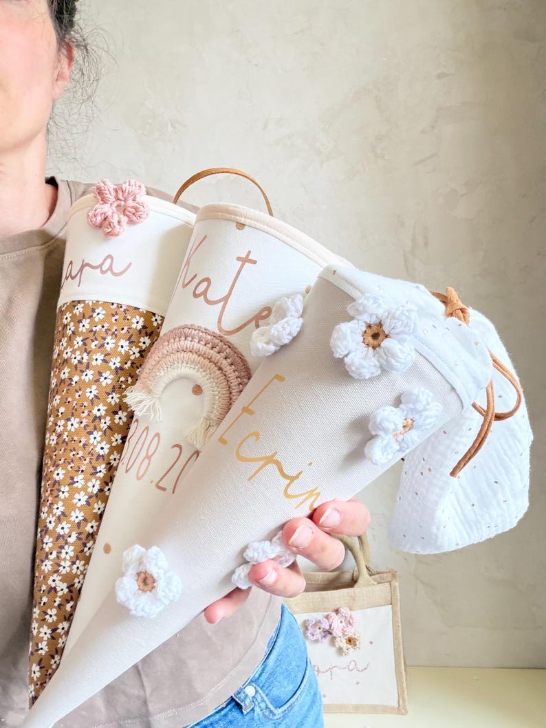 May include: Three cone-shaped fabric gift bags featuring personalized names and dates in script font. The bags are decorated with crocheted flowers and a small rainbow applique. One bag has a brown floral pattern, while others are cream-colored fabric.