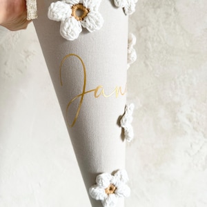 May include: A beige cone-shaped gift container featuring white crocheted flowers with tan centers attached to the exterior. The name Jan is printed in gold cursive lettering on the side. A white fabric trim with a gold metallic ribbon is at the top.