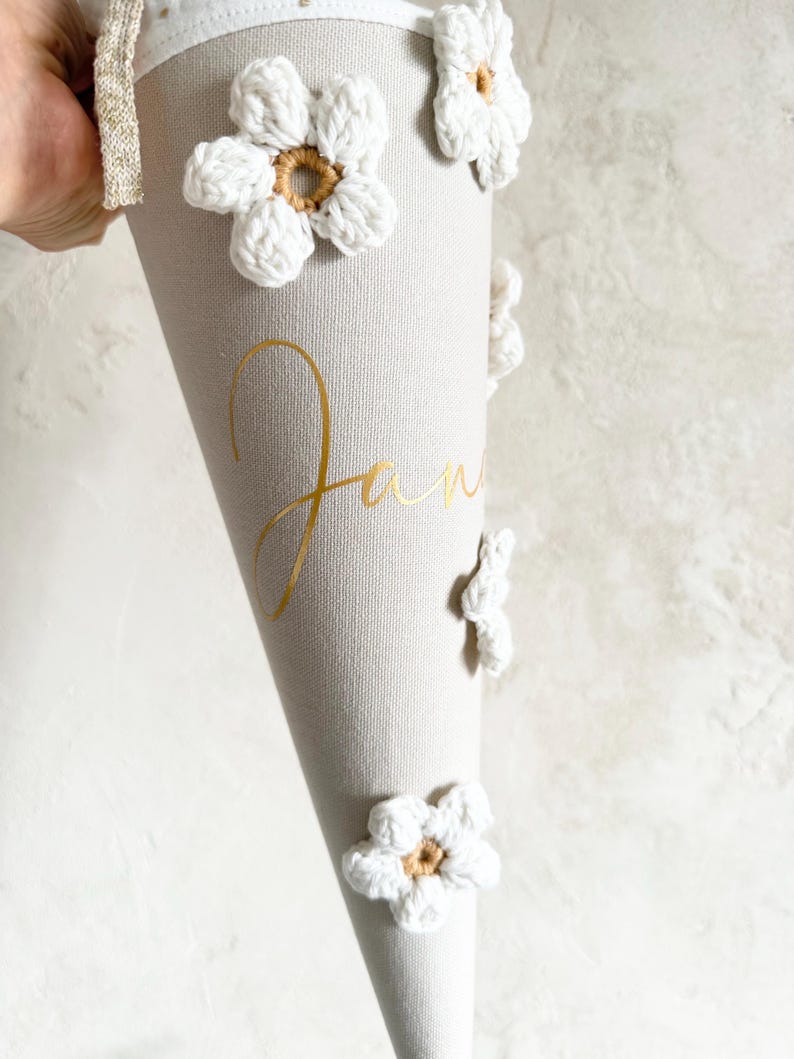 May include: A beige cone-shaped gift container featuring white crocheted flowers with tan centers attached to the exterior. The name Jan is printed in gold cursive lettering on the side. A white fabric trim with a gold metallic ribbon is at the top.