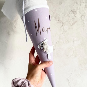 Puede incluir: Una bolsa de regalo cónica de color lavanda con lunares blancos y el nombre "Mari" en letra cursiva marrón oscuro. La parte superior tiene un cierre de tela blanca atado con un cordón gris claro. Incluye una aplicación de caballo bordado blanco.