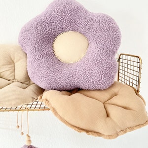 May include: A purple plush flower-shaped cushion with a cream-coloured centre. The cushion is sitting on a tan cushion with a gold metal shelf behind it.