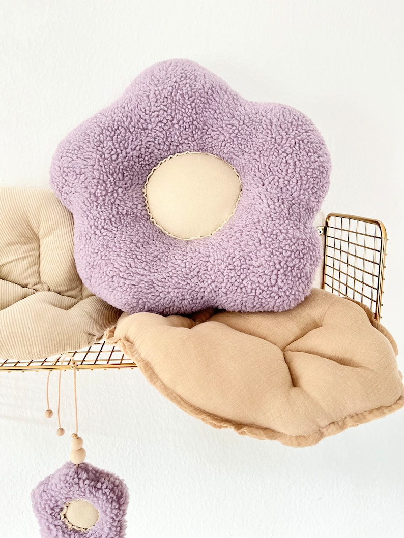 May include: A purple plush flower-shaped cushion with a cream-coloured centre. The cushion is sitting on a tan cushion with a gold metal shelf behind it.