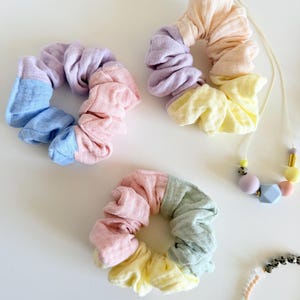 Op de afbeelding: Drie stoffen haarelastiekjes in pastelkleurige color-block patronen van blauw, roze, lavendel, geel en saliegroen, vergezeld door een ketting met geometrische siliconen kralen en een fijn armbandje met kleine gespikkelde kralen.