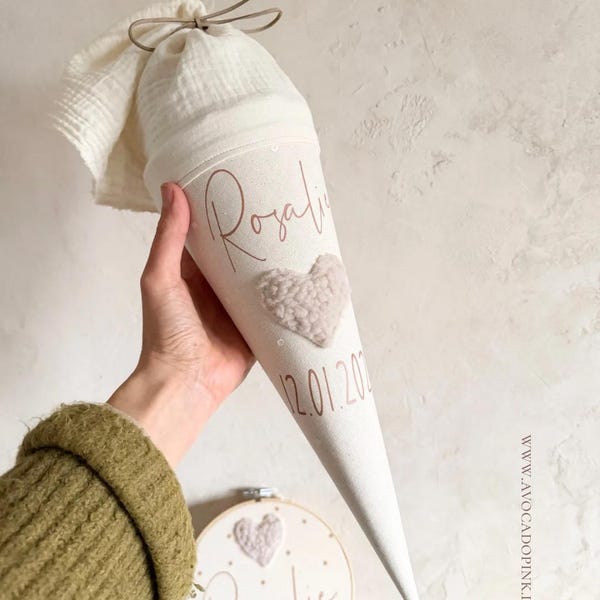 Può includere: Un cono in tessuto color crema con una parte superiore in mussola legata con un cordino. Il cono presenta il nome Rosalie e la data 12.01.2024 in corsivo marrone, con un cuore beige testurizzato. Sotto, un telaio da ricamo rotondo mostra il nome.