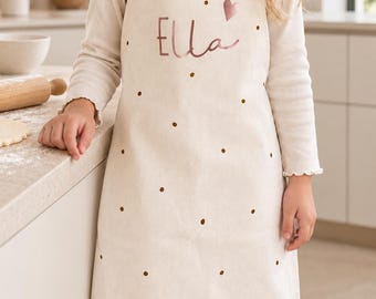 May include: A cream-colored apron featuring a repeating pattern of small brown polka dots. The front of the apron displays the name Ella in a metallic rose gold script font, accompanied by a small rose gold heart icon above the name.