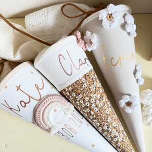 May include: Three cone-shaped fabric gift bags with drawstring closures. One features a crocheted rainbow and the name Kate with the date 3.08.2024. Another has a floral pattern and the name Clara. The third is beige with white crocheted flowers and the name Erin.