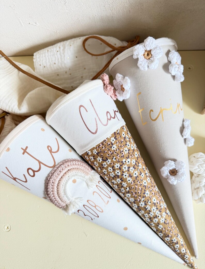 May include: Three cone-shaped fabric gift bags with drawstring closures. One features a crocheted rainbow and the name Kate with the date 3.08.2024. Another has a floral pattern and the name Clara. The third is beige with white crocheted flowers and the name Erin.
