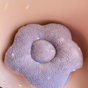 May include: A plush purple flower-shaped cushion with a raised centre. The cushion is soft and fluffy, perfect for cuddling.