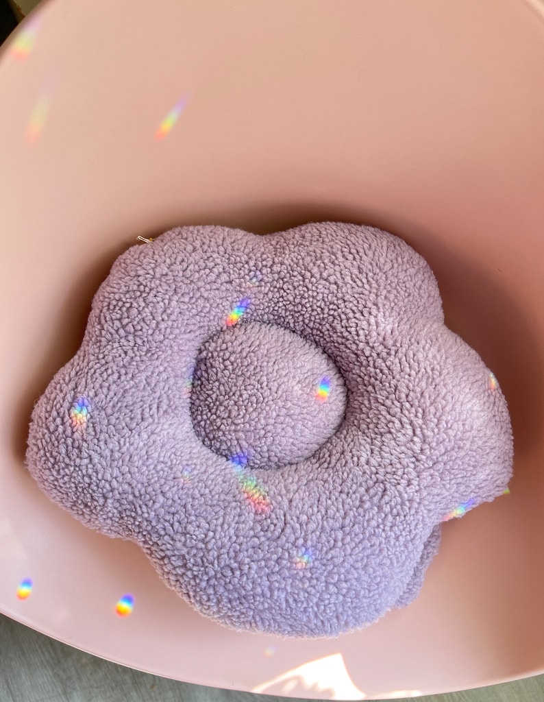 May include: A plush purple flower-shaped cushion with a raised centre. The cushion is soft and fluffy, perfect for cuddling.