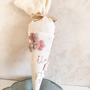 May include: A cream-colored, cone-shaped school bag with the name "Lotta" printed in rose gold. The bag is decorated with crocheted flowers in shades of pink, white, and gray. The top is tied with a beige ribbon and filled with a cream-colored fabric.