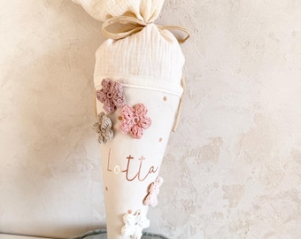 May include: A cream-colored, cone-shaped school bag with the name "Lotta" printed in rose gold. The bag is decorated with crocheted flowers in shades of pink, white, and gray. The top is tied with a beige ribbon and filled with a cream-colored fabric.