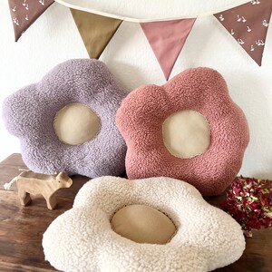 May include: Three plush flower-shaped cushions in different colours: purple, pink, and white. The cushions have a round centre and are made of soft, fluffy material.