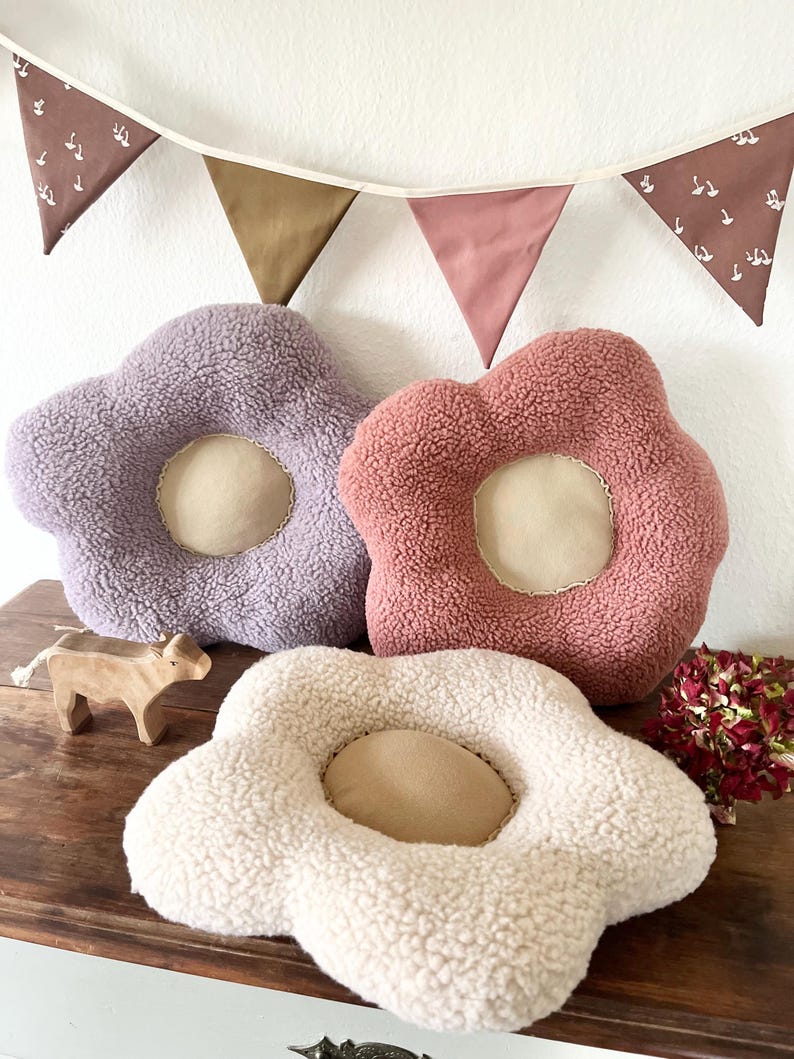 May include: Three plush flower-shaped cushions in different colours: purple, pink, and white. The cushions have a round centre and are made of soft, fluffy material.
