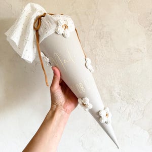 May include: A light gray cone-shaped gift bag featuring white crocheted flower appliqu&eacute;s, a white muslin fabric top with gold dots, and a brown drawstring. The bag is personalized with the name Malia and a date printed in a thin white font.