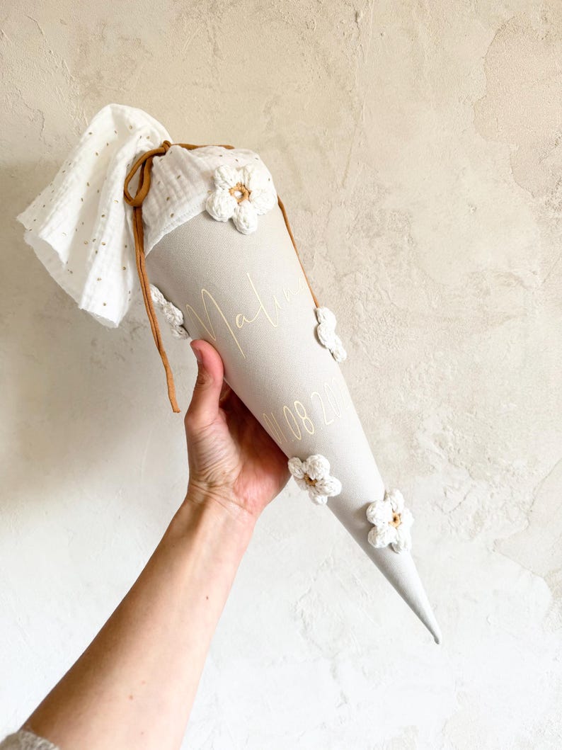 May include: A light gray cone-shaped gift bag featuring white crocheted flower appliqu&eacute;s, a white muslin fabric top with gold dots, and a brown drawstring. The bag is personalized with the name Malia and a date printed in a thin white font.