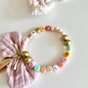 Pu&ograve; includere: Un braccialetto di perline con la parola "SCHULKIND" scritta con perline colorate. Il braccialetto presenta perline dorate e rosa. Un fiocco rosa e una forcina sono sullo sfondo.