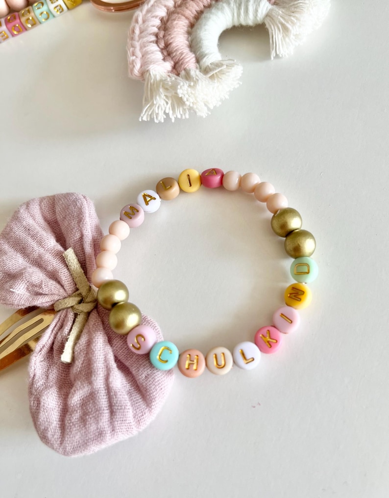 Pu&ograve; includere: Un braccialetto di perline con la parola "SCHULKIND" scritta con perline colorate. Il braccialetto presenta perline dorate e rosa. Un fiocco rosa e una forcina sono sullo sfondo.