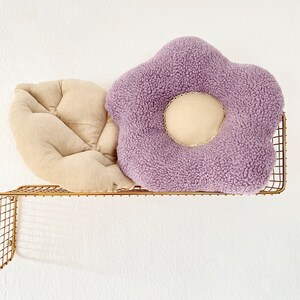 May include: Two decorative cushions on a gold metal shelf. One cushion is a light beige corduroy with a quilted design. The other cushion is a purple plush flower shape with a beige centre.