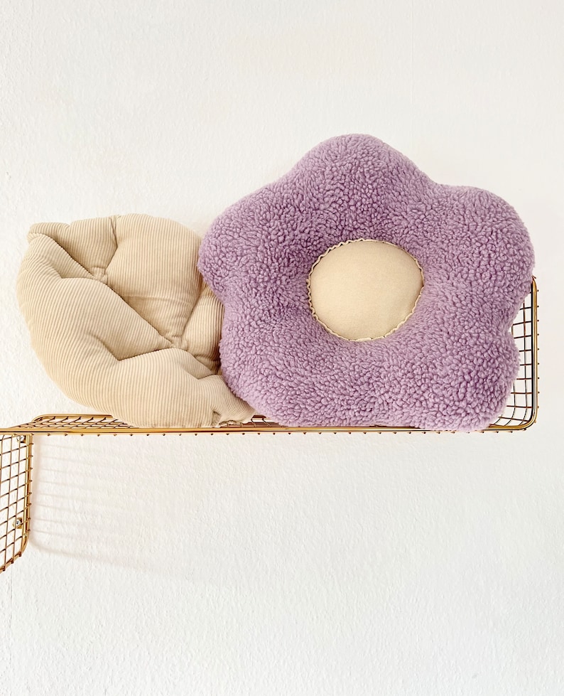 May include: Two decorative cushions on a gold metal shelf. One cushion is a light beige corduroy with a quilted design. The other cushion is a purple plush flower shape with a beige centre.