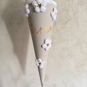 May include: A beige cone-shaped school gift bag featuring the name Beren in gold script lettering. The exterior is decorated with five white crocheted flowers with brown centers. A white fabric top with gold dots and a gold ribbon tie completes it.