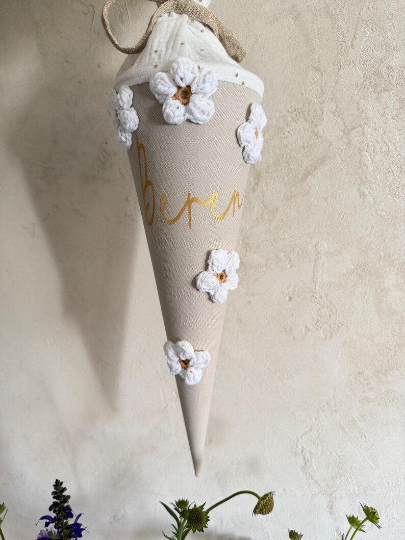 May include: A beige cone-shaped school gift bag featuring the name Beren in gold script lettering. The exterior is decorated with five white crocheted flowers with brown centers. A white fabric top with gold dots and a gold ribbon tie completes it.