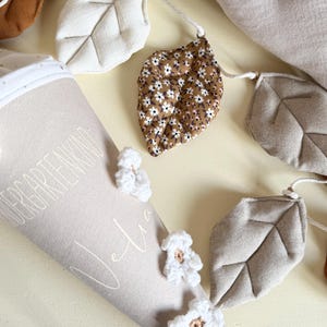 May include: A beige fabric cone with the text "KINDERGARTENKIND" and "Julia" printed on it, decorated with white crocheted flowers. Beside it is a garland featuring fabric leaves in solid beige, brown, and a brown floral pattern.