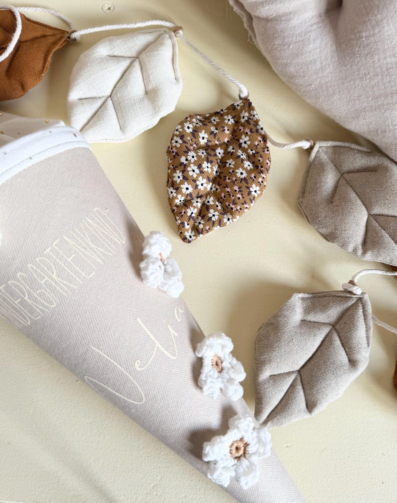 May include: A beige fabric cone with the text "KINDERGARTENKIND" and "Julia" printed on it, decorated with white crocheted flowers. Beside it is a garland featuring fabric leaves in solid beige, brown, and a brown floral pattern.