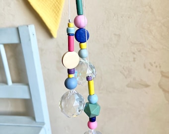 May include: Two crystal suncatchers with colorful wooden beads hanging from white strings. The beads are in shades of pink, blue, green, yellow, and purple. The suncatchers are hanging in front of a light brown wall with a yellow and pink fabric banner in the background.