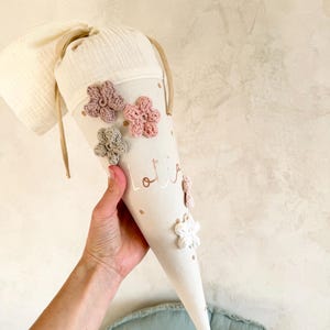 May include: A beige school cone decorated with crocheted flowers in pink, white, and green. The cone has the name "Lotta" printed on it in gold. The cone is held by a person.