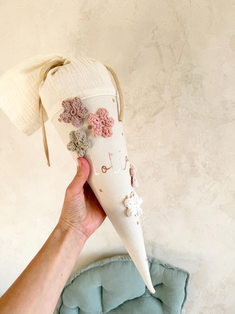 May include: A beige school cone decorated with crocheted flowers in pink, white, and green. The cone has the name "Lotta" printed on it in gold. The cone is held by a person.